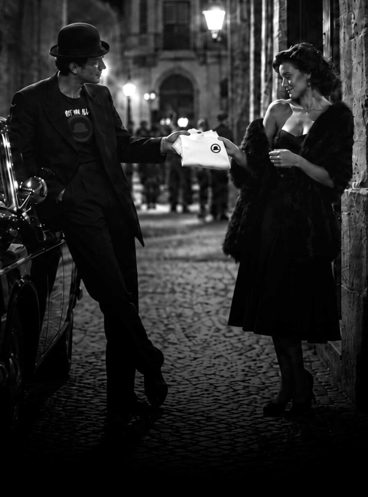 Uomo con bombetta porge una t-shirt Milord a una donna in una via di notte